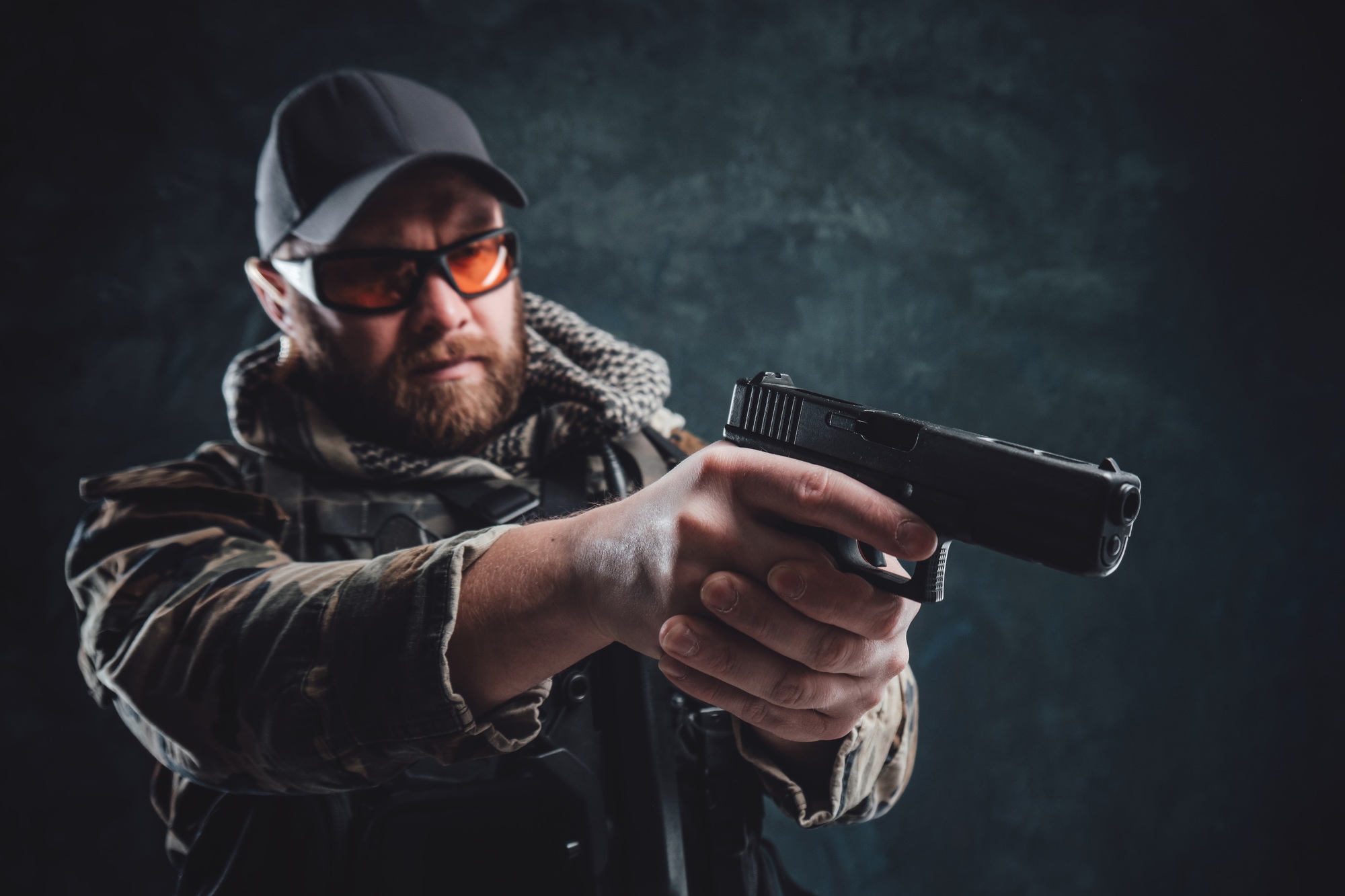 Army soldier with a cap and glasses aiming a handgun in dark background